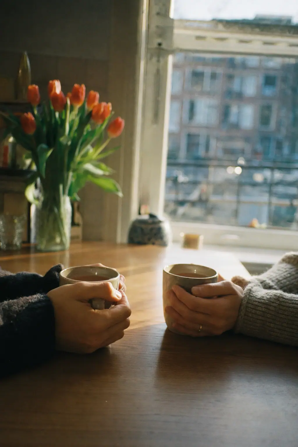 Two hands around mugs of tea at a kitchen table by the window