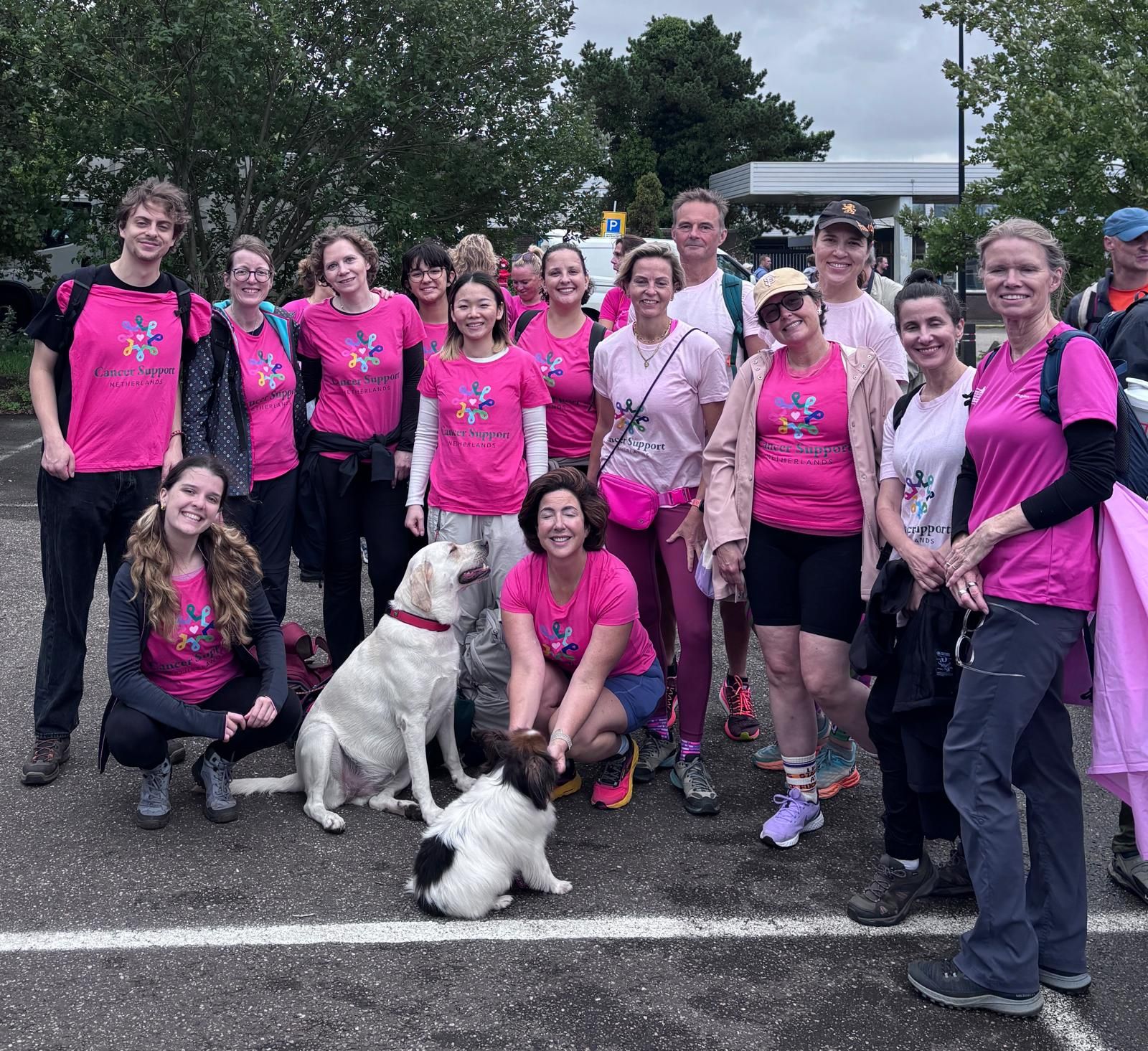 A large group of Cancer Support Netherlands members and a dog, all in pink CSN T-shirts, smiling together in a city square